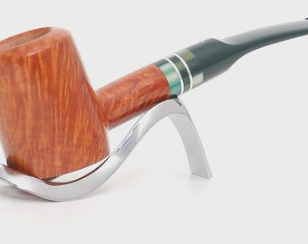 May include: A tobacco pipe with a polished light brown wooden bowl and a dark green stem. The stem features a decorative band with silver and green accents. The pipe rests on a curved, silver-coloured metal stand with a smooth, reflective finish.