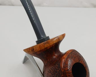 May include: A handcrafted tobacco pipe with a dark grey stem and a wooden bowl. The bowl is a rich brown colour with a textured surface and a smooth, polished finish. The pipe rests on a silver-coloured stand.