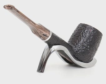 May include: A tobacco pipe with a dark brown, textured bowl and a marbled brown and tan stem. The pipe is supported by a polished silver-coloured metal stand that cradles the base of the bowl and the shank, featuring a sleek, curved design.