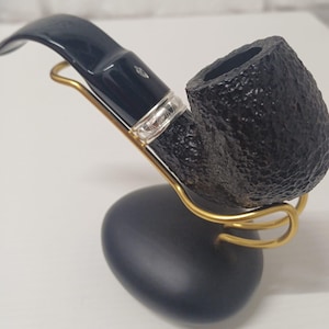 Peut inclure: Une pipe à tabac noire avec un foyer texturé et un tuyau incurvé, posée sur un support doré. La tige comporte une bague argentée et un petit détail en forme de losange. Le support est soutenu par une base bleu foncé.