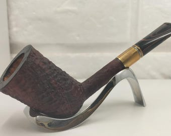 May include: A tobacco pipe with a dark brown, textured bowl and stem, featuring a gold-colored metal band connecting to a black and brown marbled acrylic mouthpiece. The pipe rests on a curved, polished silver-colored metal stand.