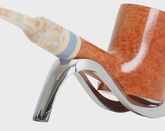 May include: A tobacco pipe with a polished light brown wooden bowl and a marbled cream-coloured stem. The stem features a light blue band near the bowl. The pipe rests on a curved, silver-toned metal stand with a smooth, reflective finish.