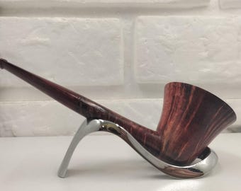 May include: A tobacco pipe with a polished dark brown wood grain finish and a long, slender stem, resting securely on a curved, silver-toned metal stand. The pipe features a deep, wide bowl with visible natural wood patterns and a smooth texture.