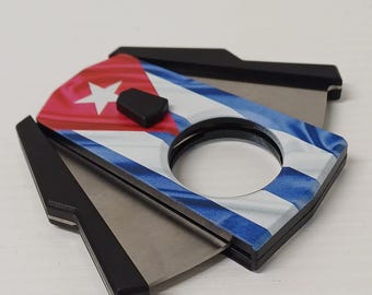 May include: A cigar cutter featuring the Cuban flag. The design incorporates the red, white, and blue colours of the flag, with a white star. The blades are silver, and the handle is black.
