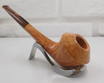 May include: A wooden tobacco pipe with a curved stem and bowl, resting on a silver-colored stand. The pipe features a light brown finish with darker wood accents on the stem. The bowl has a large opening.