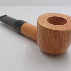 May include: A light-colored, wooden tobacco pipe with a rounded bowl and a dark brown stem. The pipe has a classic design, with a smooth, polished finish. The bowl is cylindrical and has a large opening. The stem is curved and tapers to a mouthpiece.
