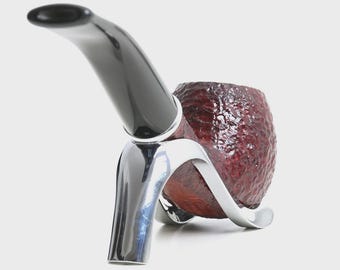 May include: A tobacco pipe with a dark brown, textured bowl and a smooth, curved black stem. The pipe is supported by a polished, silver-coloured metal stand that features a tripod-like base, holding the bowl securely in a horizontal position.