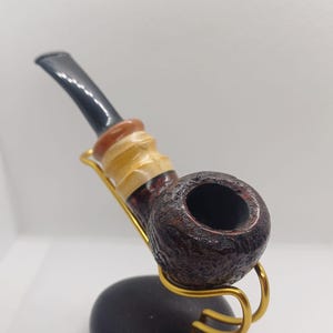 May include: A dark brown wooden pipe with a black stem and a multi-colored band around the bowl. The pipe is sitting on a gold metal stand.