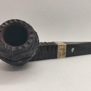 May include: A dark brown tobacco pipe with a textured bowl and a smooth, black stem. The pipe features a silver band with an emblem and the letter "φ". The bowl has a carved, organic design.