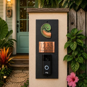 May include: A rectangular black plaque mounted on a beige pillar featuring a green and copper nautilus shell emblem, a copper address plate with the text 1233 Imperial Drive, and a black Ring video doorbell with a glowing blue circular button.