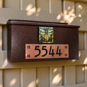 May include: A brown mailbox with a textured finish, featuring a stained glass dragonfly design and a copper address plate with the number 5544. The mailbox is mounted on a light-colored wooden wall.