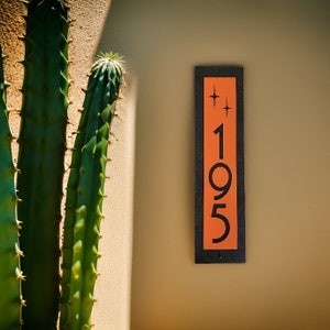 May include: A black and orange house number sign with the number 195 and two small stars above the number.