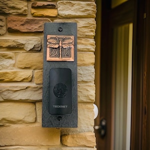 May include: A black doorbell with a copper dragonfly design on a stone wall. The doorbell has the text "TECKNET" on it.