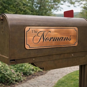 May include: A bronze-colored metal mailbox featuring a rectangular copper-toned plaque with a decorative black border. The text "The Normans" is engraved in a black script font on the plaque. A small red flag is attached to the top right side.