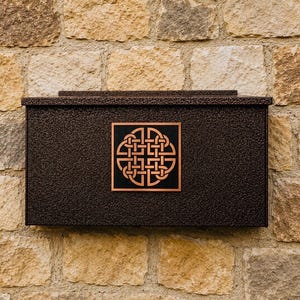 May include: A rectangular, dark bronze-colored metal mailbox with a textured finish. The front panel features a centered square plaque displaying a copper-toned Celtic knot design set against a black background, mounted on a light-colored stone wall.