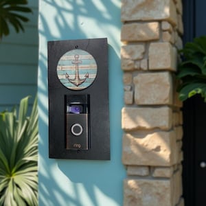 May include: A black rectangular doorbell mount with a circular anchor design. The anchor is brown and set against a blue and white striped background. A Ring doorbell is installed below the anchor design.