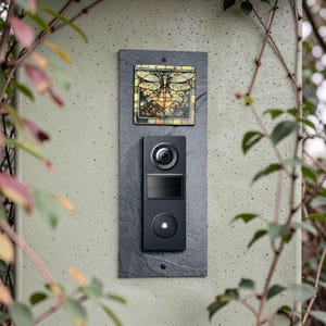 May include: A black smart doorbell system mounted on a dark gray slate board. The system includes a camera, a speaker, and a doorbell button. Above the doorbell is a decorative tile with a dragonfly design in shades of yellow, orange, and green.