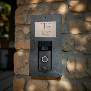 May include: A doorbell system mounted on a dark gray rectangular slate. The system includes a bronze-colored doorbell with a camera and the word "ring" at the bottom. Above the doorbell is a rectangular plaque with the number "119" and the words "Navtem Place".