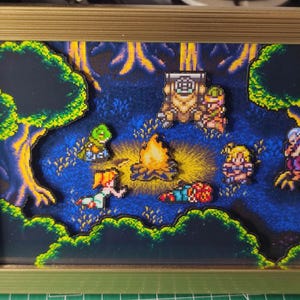 Può includere: Una scena di pixel art incorniciata che raffigura un falò circondato da personaggi in un ambiente forestale. L'opera presenta una cornice dorata, uno sfondo di foresta blu e verde e diverse figure pixelate. Il falò è al centro.
