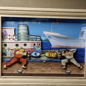 May include: A 3D shadow box art piece featuring a Street Fighter scene with Ken and Ryu in a fighting stance on a dock. The background displays a ship with the text CAPCOM. The characters are layered to create depth within a light wood frame.