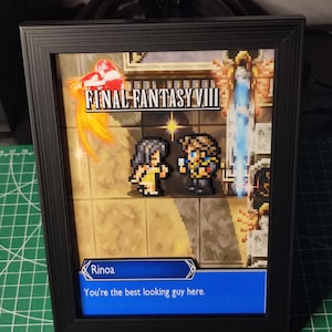 Puede incluir: Obra de arte enmarcada en negro con personajes de píxeles de Final Fantasy VIII. La imagen incluye el título del juego y un cuadro de diálogo con el texto "You're the best looking guy here." El marco es negro.