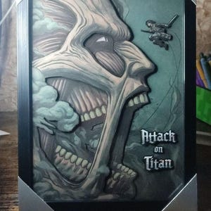Op de afbeelding: Ingelijste illustratie van een Titan uit "Attack on Titan". Het kunstwerk gebruikt grijstinten en groen, met de zijkant van het gezicht van de Titan. De titel "Attack on Titan" staat eronder.