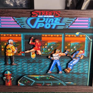 Può includere: Opera d'arte incorniciata raffigurante una scena pixelata del videogioco "Streets of Rage". L'immagine mostra personaggi in azione, un idrante e il logo del gioco. La cornice è di colore marrone scuro.