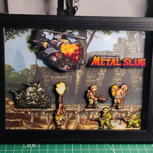 Pode incluir: Obra de arte emoldurada com personagens e veículos pixelizados do videogame Metal Slug. A cena inclui um helicóptero em chamas, soldados e um tanque. O título "Metal Slug" é exibido em destaque.