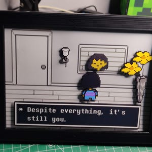 May include: Framed pixel art scene from the video game Undertale. The black frame surrounds a grayscale interior with a character and text that reads "Despite everything, it's still you." Yellow flowers and a door are also visible.