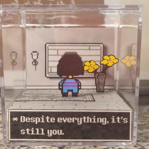 Può includere: Un diorama in cubo acrilico trasparente con un personaggio in pixel art dai capelli scuri che indossa una maglietta a righe blu e viola. La scena include una parete grigia con una cornice rettangolare, un vaso con fiori gialli e una casella di testo che recita: * Despite everything, it's still you.