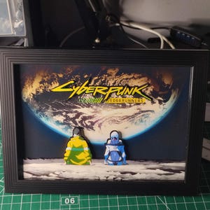 Peut inclure: Œuvre encadrée avec le logo "Cyberpunk: Edgerunners" et deux personnages, sur fond de la Terre. Le cadre noir entoure l'image, qui présente une palette de couleurs vives.