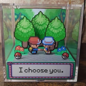 Può includere: Diorama in pixel art in una scatola di acrilico trasparente. Raffigura due personaggi che si stringono la mano in una scena di foresta. Include alberi pixelati, fiori e due Poké Ball. Il testo "I choose you." è presente.