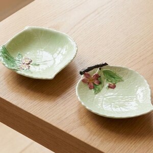 May include: Two small, light green ceramic dishes shaped like leaves. Each dish features a raised, three-dimensional floral accent with pink petals, green leaves, and a dark brown stem detail. The surfaces have a glossy finish and visible veins.