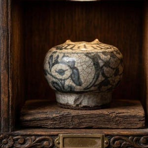 May include: A small, round ceramic jar with a crackled off-white glaze and dark blue floral patterns. The jar features a wide, rounded body, a short neck, and a slightly flared rim, resting on a thick, dark wooden block within a wooden cabinet.