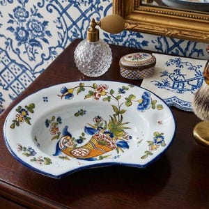May include: A white ceramic shaving bowl with a blue rim and a hand-painted floral design featuring a central cornucopia filled with flowers. The bowl has a distinct curved cutout on one side and is decorated with small blue, yellow, and green blossoms.