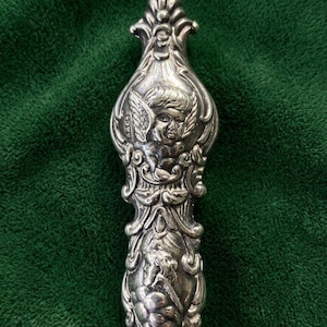 May include: A silver-toned metal handle featuring an ornate relief design. The top section displays a winged cherub figure, while the lower portion shows a secondary figure in profile. The surface has intricate scrollwork and decorative detailing.