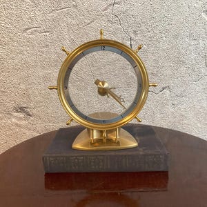 May include: A gold-toned ship wheel clock with a transparent face and metallic hands. The clock features a circular frame with spokes and is mounted on a solid, triangular base. The numbers 3, 6, 9, and 12 are marked on the inner silver ring.