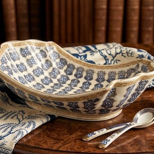 May include: An oval ceramic serving bowl with a scalloped edge, featuring a blue and white repeating lattice pattern and gold trim. Two matching ceramic spoons with blue and white floral motifs rest beside the bowl on a wooden surface.