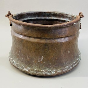 May include: A weathered, round copper pot with a dark, oxidised patina and visible signs of age. The vessel features a wide, open top, a curved body, and two metal side handles attached to the rim. The base has a distinct, textured seam.