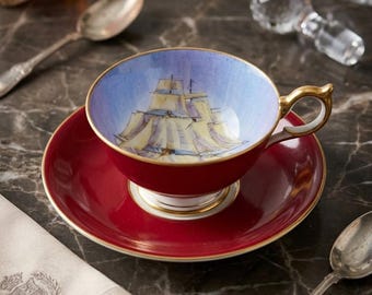 Può includere: Un set di tazza e piattino in porcellana con esterno rosso intenso e bordo dorato. L'interno della tazza mostra un'illustrazione dipinta di un veliero su uno sfondo di cielo azzurro. Il manico è color oro e di forma curva.