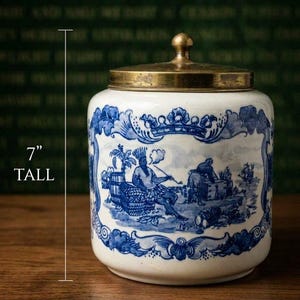 May include: A white ceramic jar with a blue transferware pattern depicting figures in a landscape, topped with a brass-coloured metal lid. The jar is 7 inches tall and 6 inches wide, featuring a decorative border and a central scene with a person smoking.