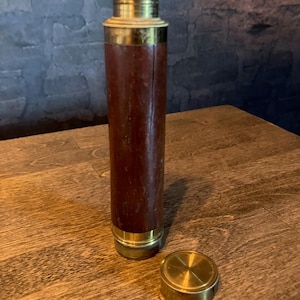 Puede incluir: Un telescopio de estilo vintage con un cuerpo de cuero marrón oscuro y detalles en latón. El telescopio cilíndrico tiene una tapa de latón extraíble al lado. Los detalles de latón incluyen la parte superior, la inferior y una banda en el medio.