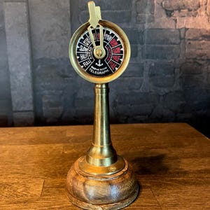 Può includere: Un telegrafo per sala macchine navale in ottone con un quadrante circolare che presenta testo nero e rosso, incluse le parole Engine Room Telegraph e un simbolo di ancora. Il dispositivo ha una leva centrale su una base in legno.