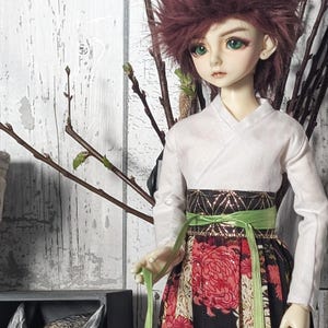 May include: A doll wearing a white kimono with a green and gold obi. The doll has long, dark red hair and green eyes. The doll is standing in front of a gray wooden background with a dragon statue in the foreground. The doll is wearing a black and red floral skirt.