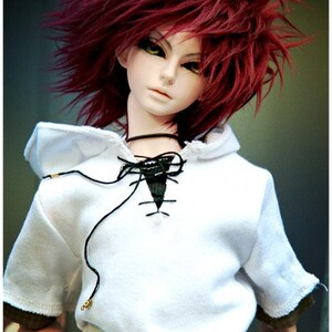 May include: A doll with long, bright red hair wearing a white hooded shirt with a black lace-up detail. The doll has a white face with green eyes and a neutral expression.