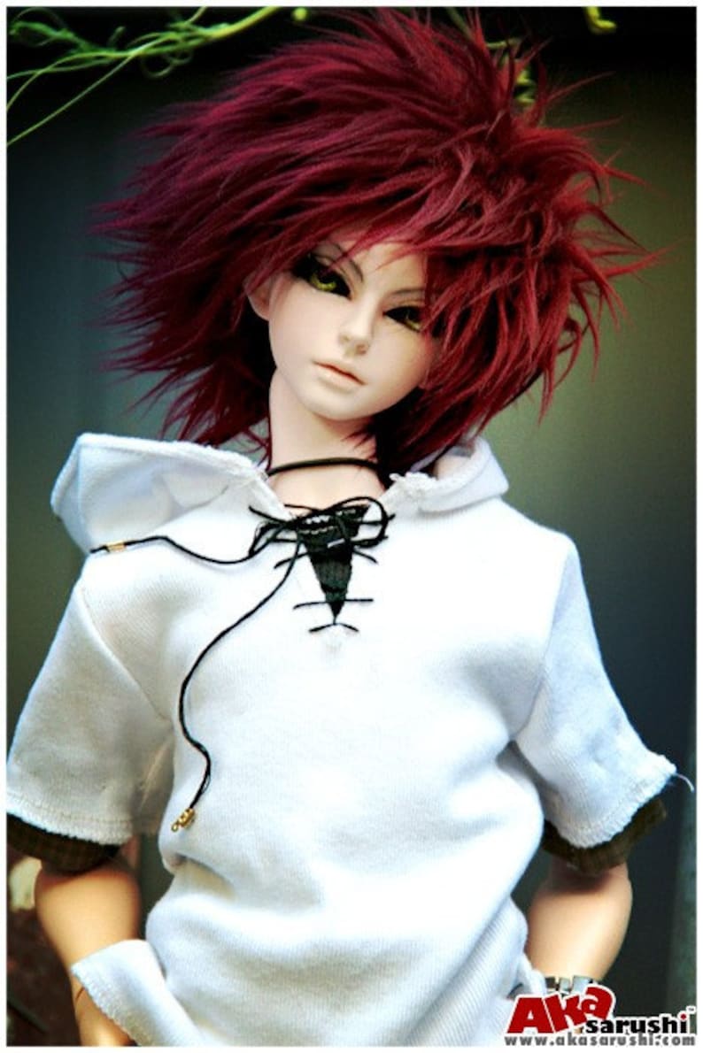 May include: A doll with long, bright red hair wearing a white hooded shirt with a black lace-up detail. The doll has a white face with green eyes and a neutral expression.