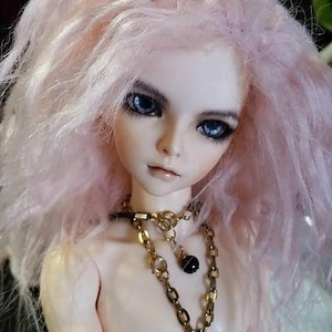 May include: A doll with long, pink hair and blue eyes. The doll is wearing a gold necklace with a spider pendant.