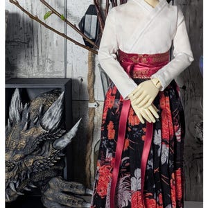 May include: A doll wearing a white kimono with a red sash and a black and red floral skirt. The doll has long, red hair and green eyes. The doll is standing in front of a wooden background.