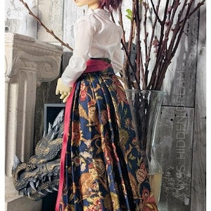 May include: A doll wearing a white shirt with a red sash and a long, pleated skirt with a floral print. The skirt features a pattern of birds, flowers, and leaves in shades of gold, red, and blue on a dark blue background.