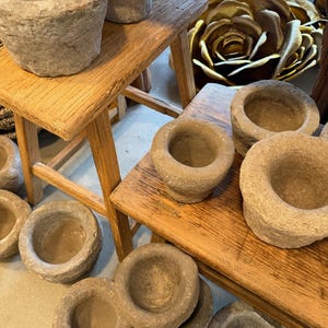 Puede incluir: Una colección de juegos de mortero y maja de piedra de varios tamaños, exhibidos sobre taburetes de madera. Los morteros son de color gris claro con una textura rugosa. Los taburetes son de madera de color claro. Grandes esculturas decorativas de rosas doradas en el fondo.